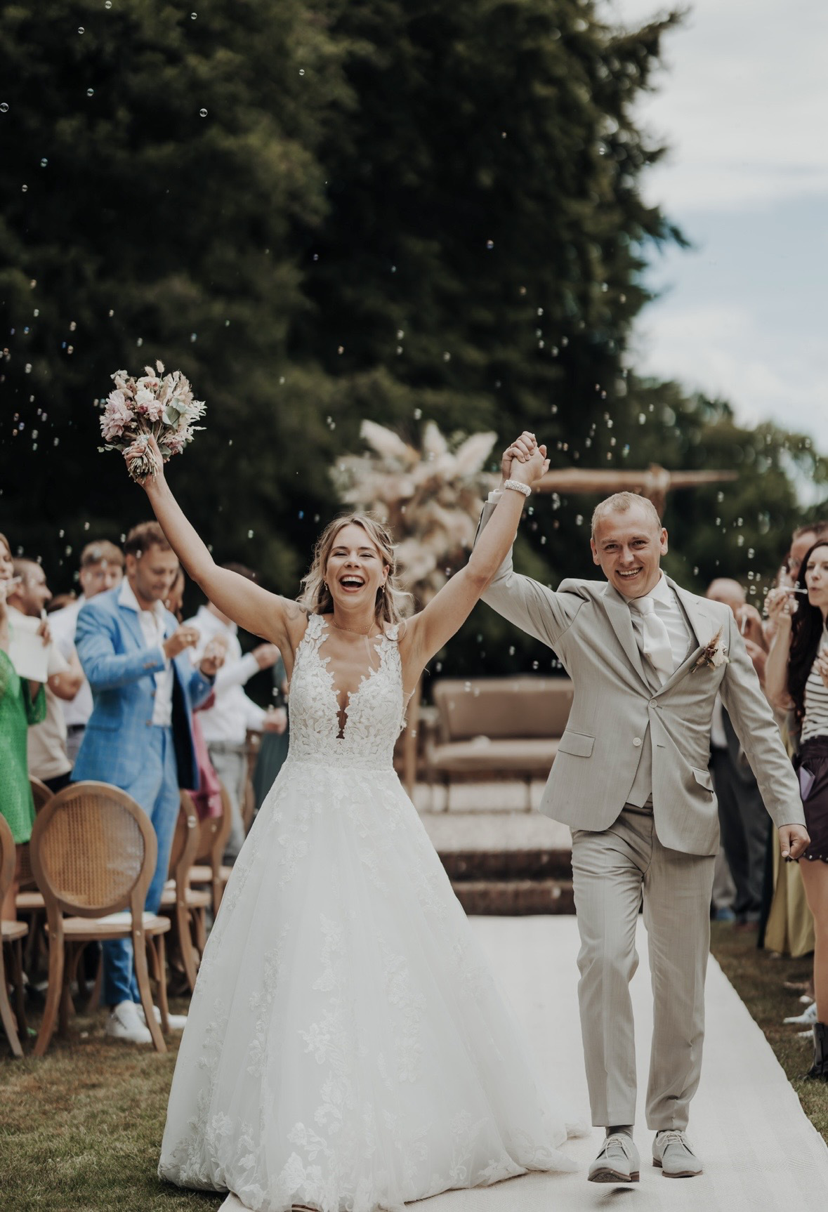 real bride sabrina veldman ehlebracht in een romantische a-lijn trouwjurk juichend samen met haar bruidegom tijdens de ceremonie met boeket omhoog en gasten op de achtergrond